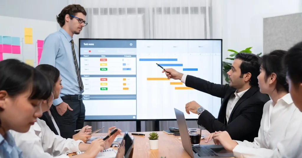 Business team discussing project timeline on screen during meeting while taking notes on laptops and tablets.